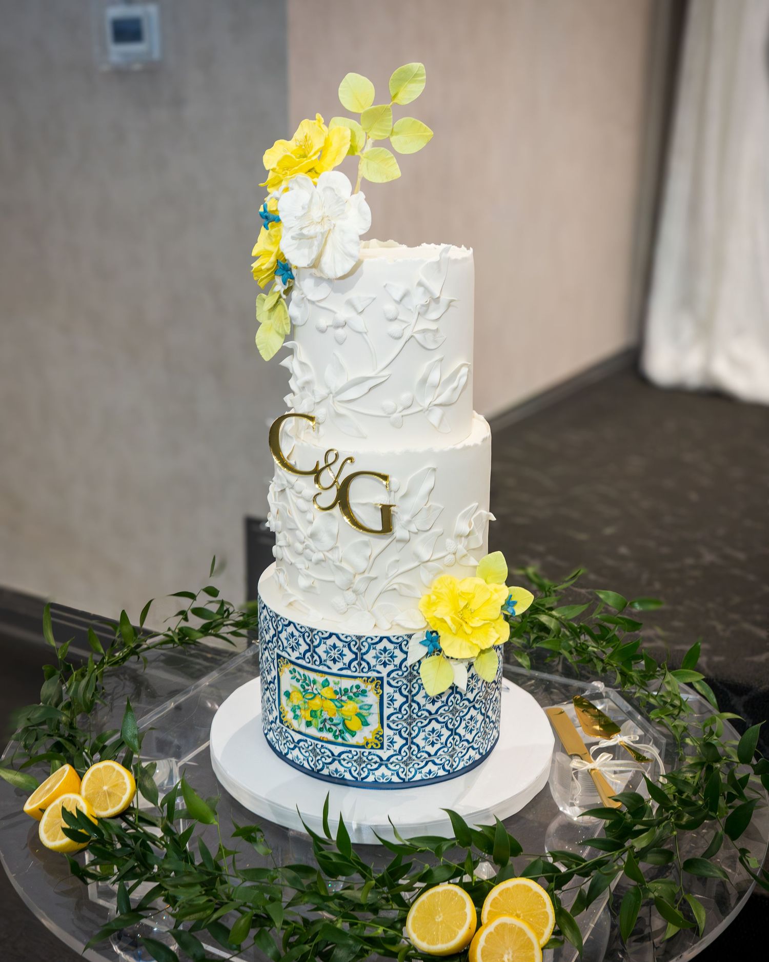 Three-tier wedding cake with blue tile design, yellow roses, white flowers, C&G monogram, surrounded by lemons and greenery.