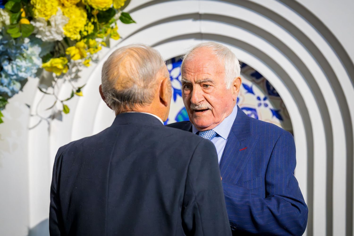 Two elderly men in suits having a conversation near a decorative floral arch with yellow and blue flowers.