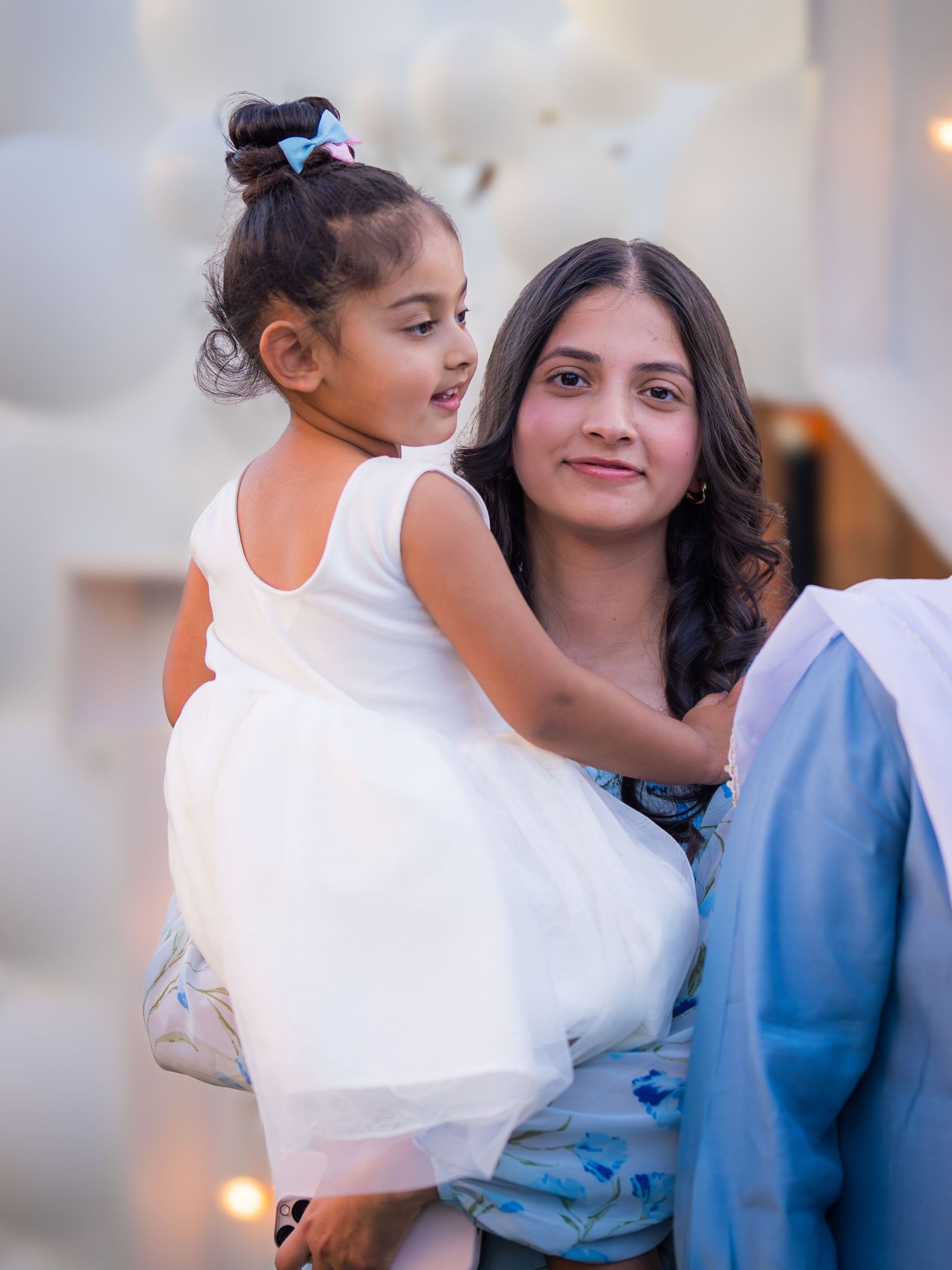 A young woman holds a little girl in a white dress at a gender reveal celebration with balloons.