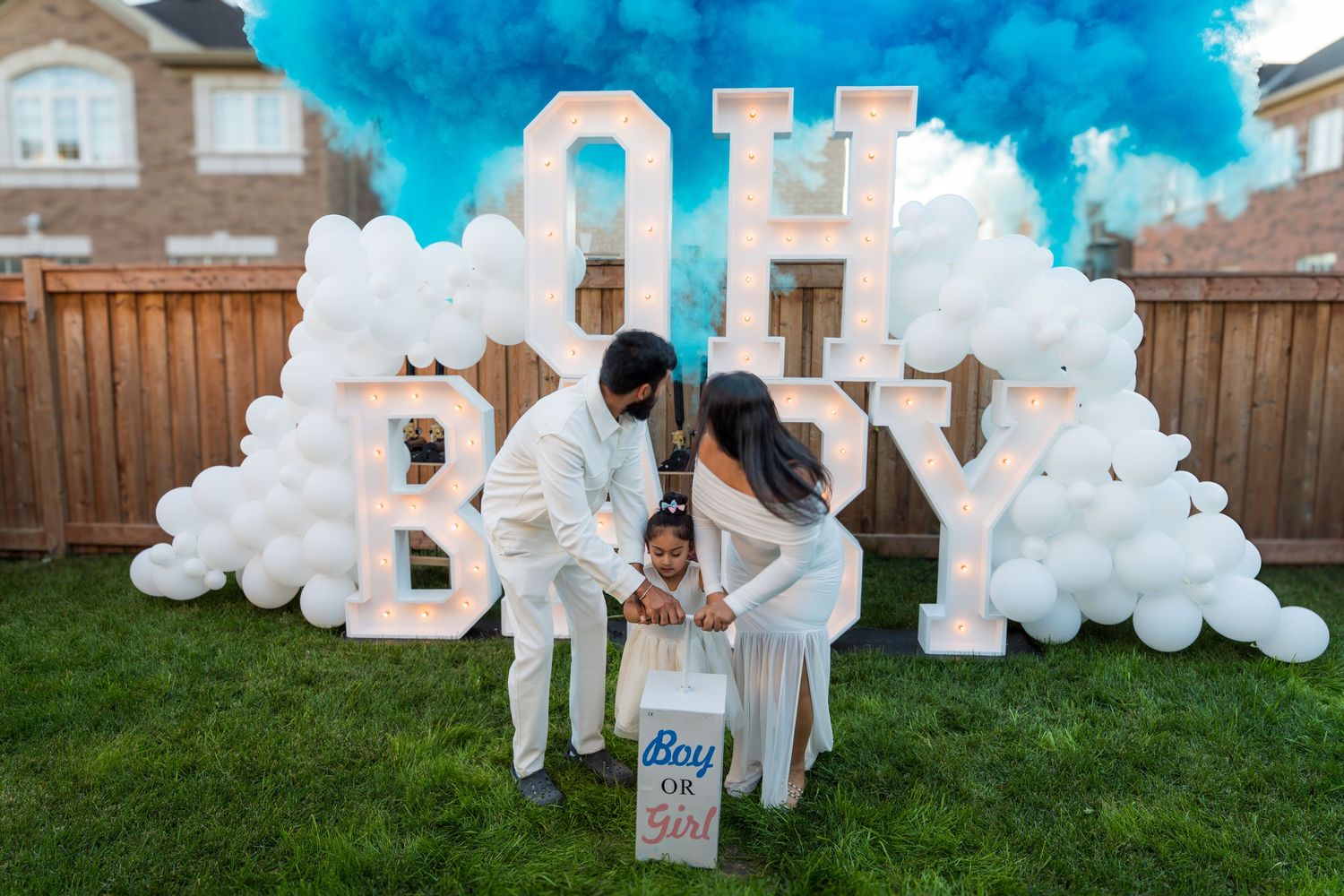 Couple with child at blue smoke gender reveal party with OH BABY marquee letters and white balloons.