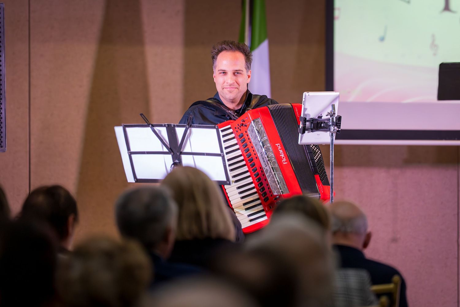 A smiling man plays a red Roland accordion on stage before an audience at an indoor event.
