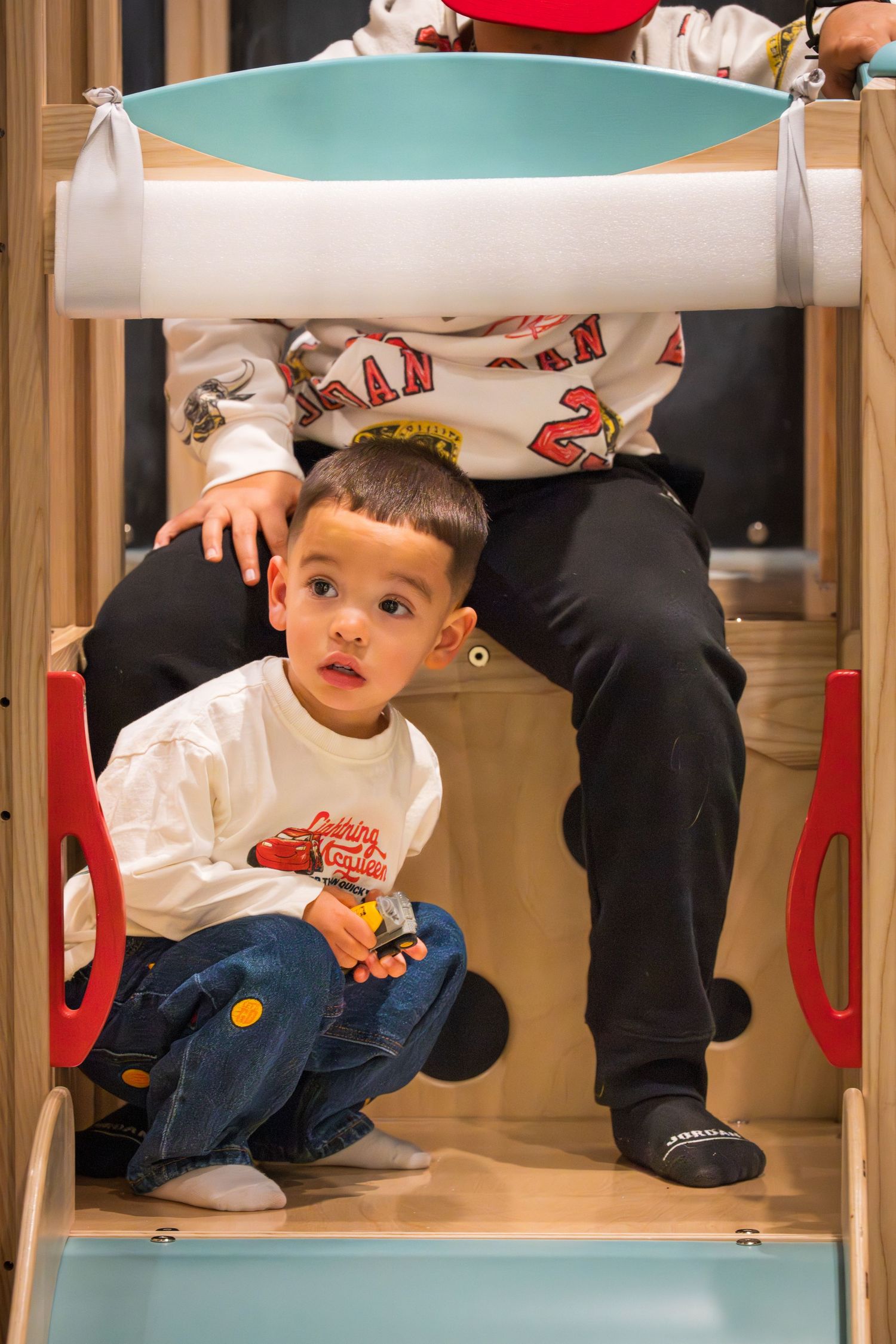 Young boy crouching on wooden playground slide indoors, wearing white shirt and jeans, holding a toy.