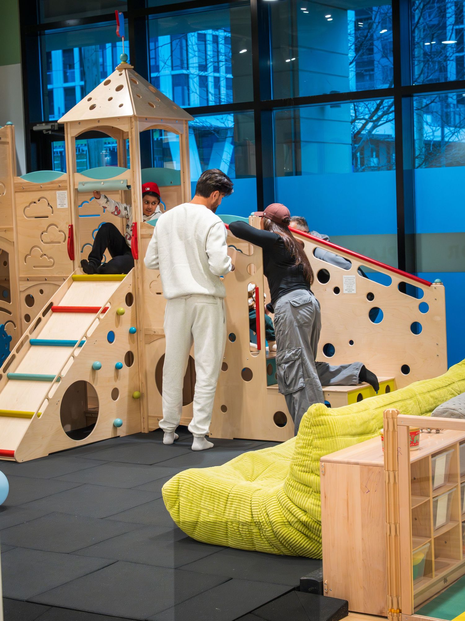 Adults assist children on a wooden indoor playground climbing structure with slides and foam seating nearby.