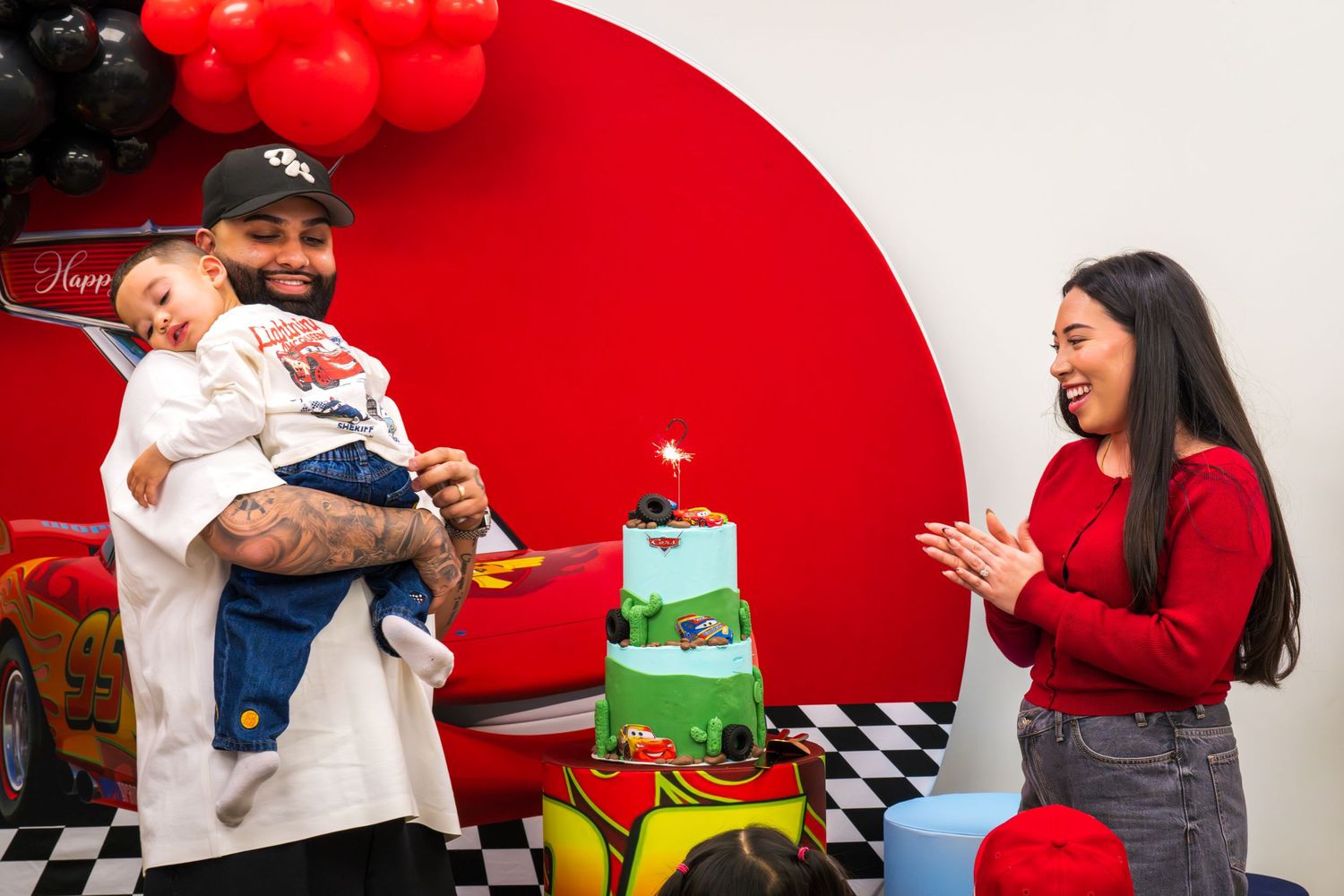 Tattooed father holding baby boy next to Disney Cars birthday cake while smiling mother claps at party.