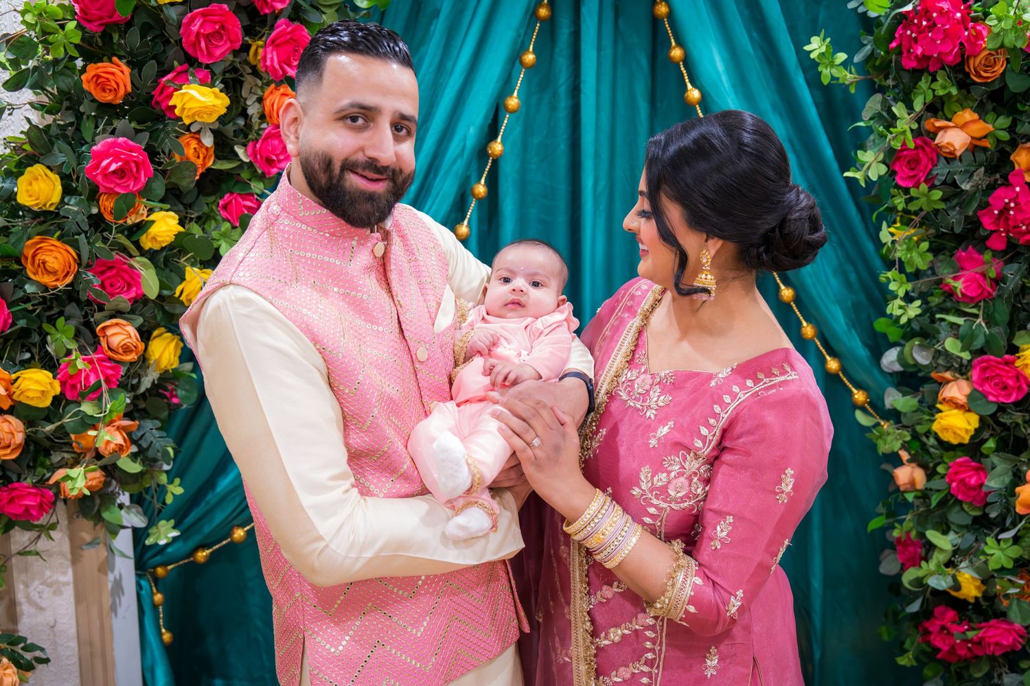 Indian couple dressed in pink ethnic wear smiling while holding their newborn baby at a celebration.