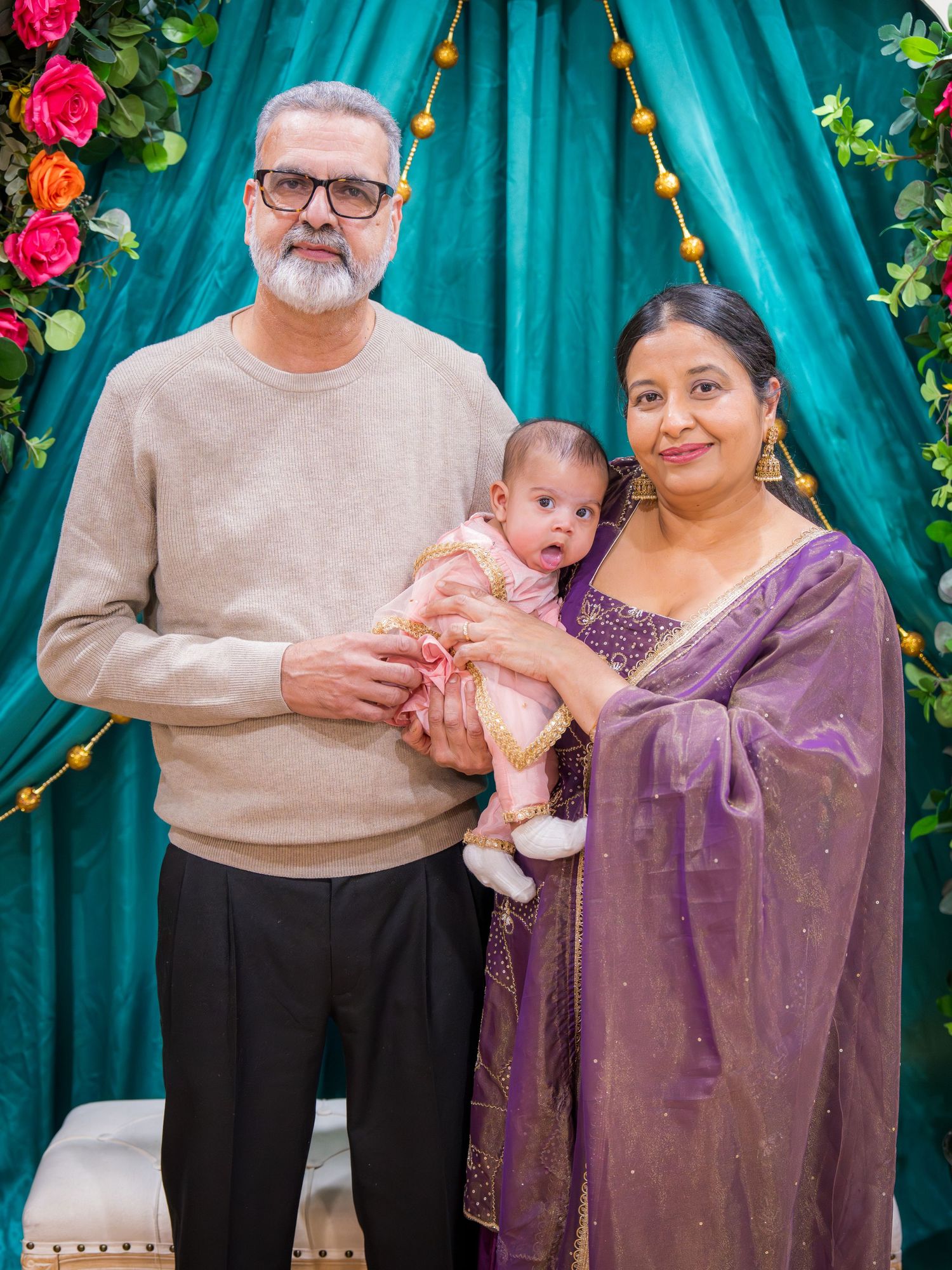Grandparents in traditional South Asian attire holding a baby at a festive celebration with teal drape backdrop.