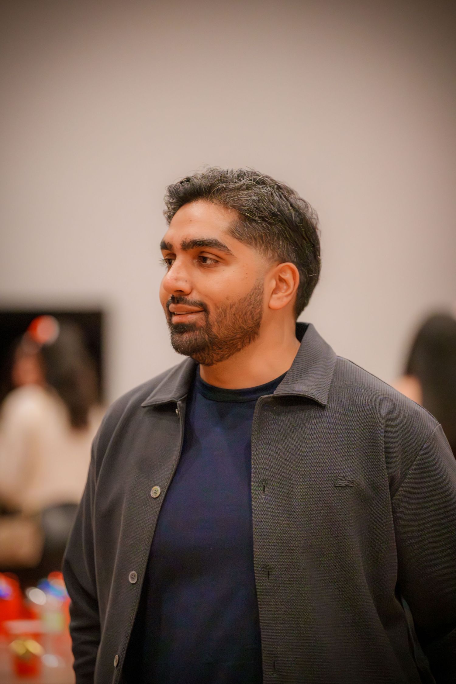 A person wearing a navy blue sweater and dark jacket smiles while looking to the side at an indoor gathering.