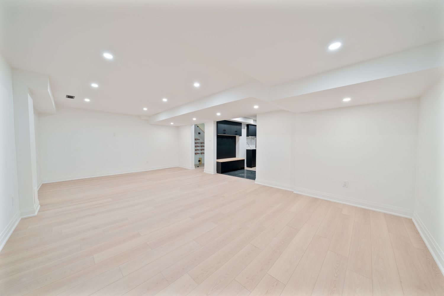 Empty basement room with light wood flooring, white walls, recessed lighting and a doorway in the distance.