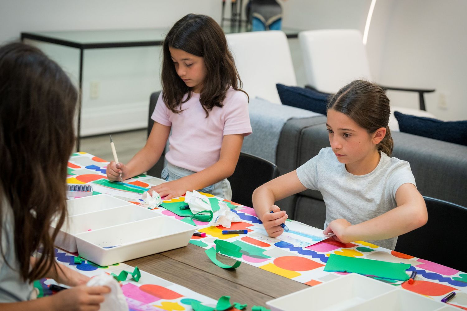 Three girls collaborate on a colorful art project at a table filled with bright paper cutouts.