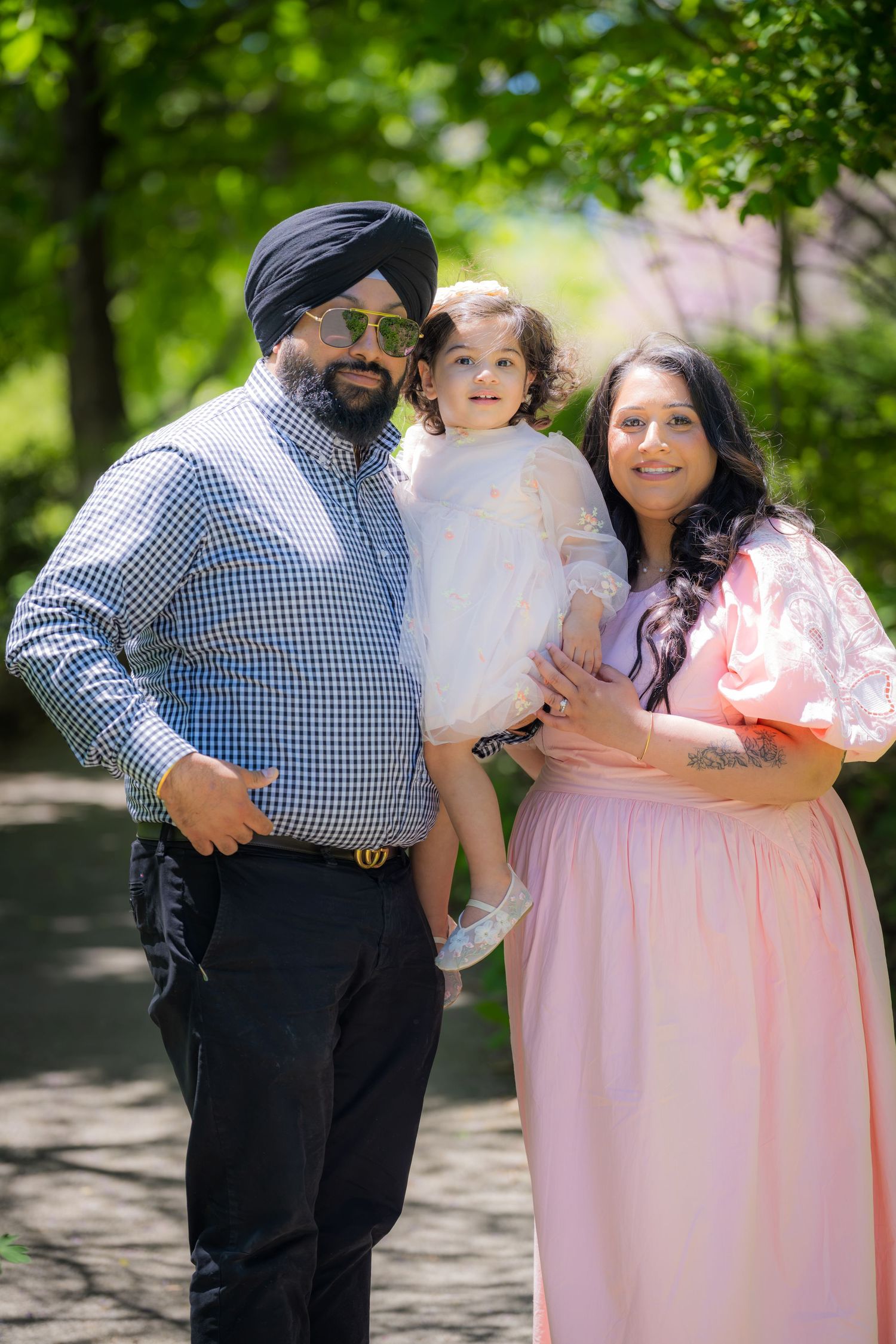 A Sikh man and woman in pink dress smile holding a baby girl in white dress outdoors.