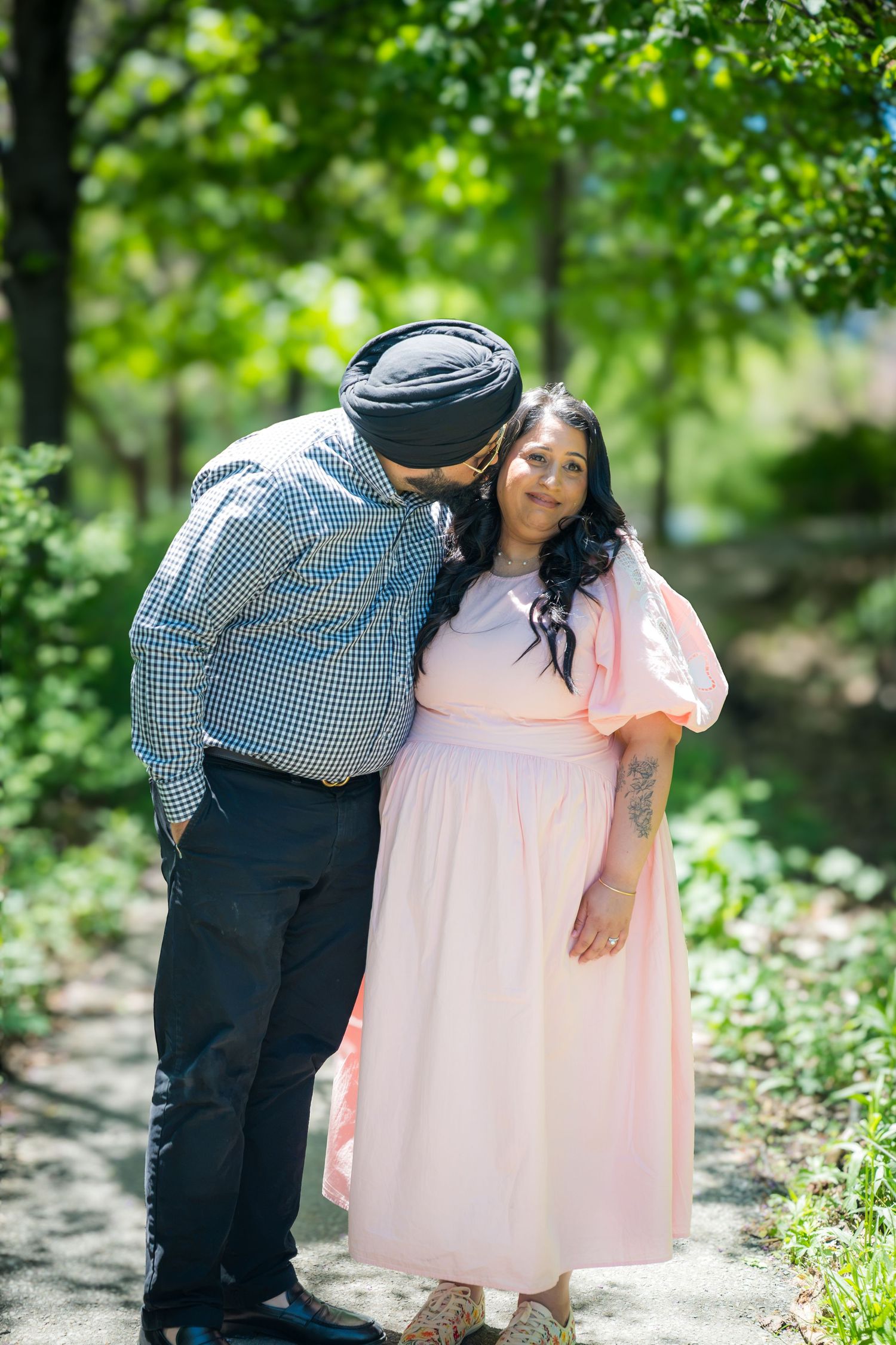 A Sikh man tenderly kisses a pregnant woman in a pink dress in a sunlit green park.