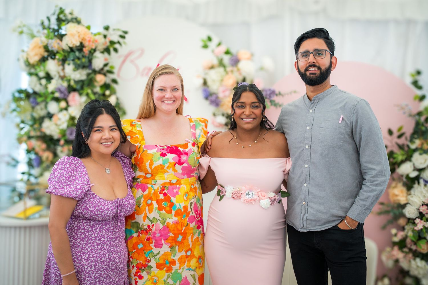 Friends posing together in front of a floral wall backdrop at a baby shower celebration.