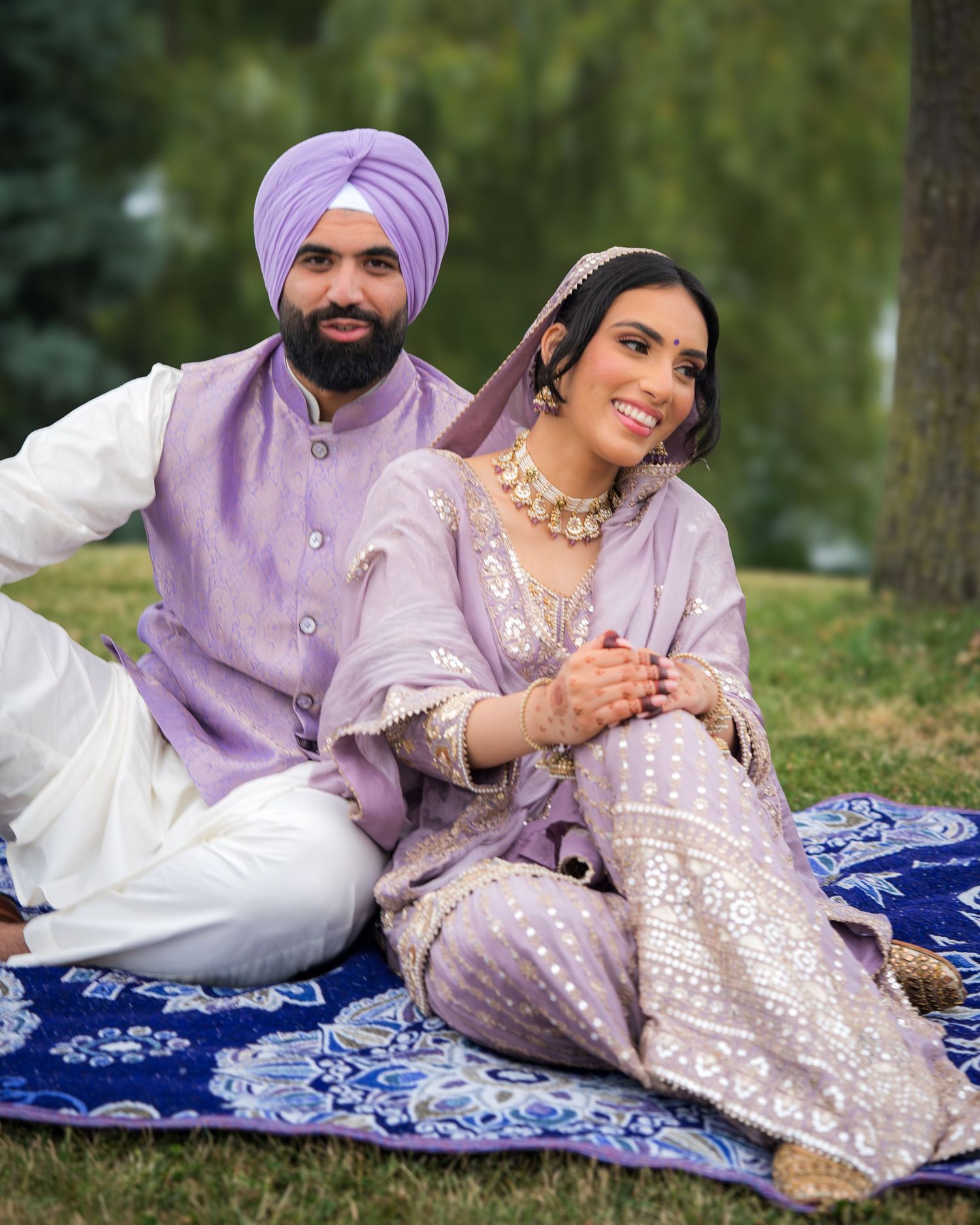 Romantic couple lounging on decorative blue blanket wearing matching lavender and white traditional attire.