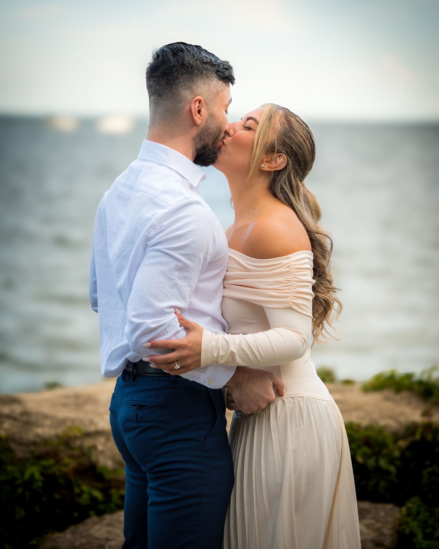 A romantic couple embraces on a rocky shoreline at sunset.