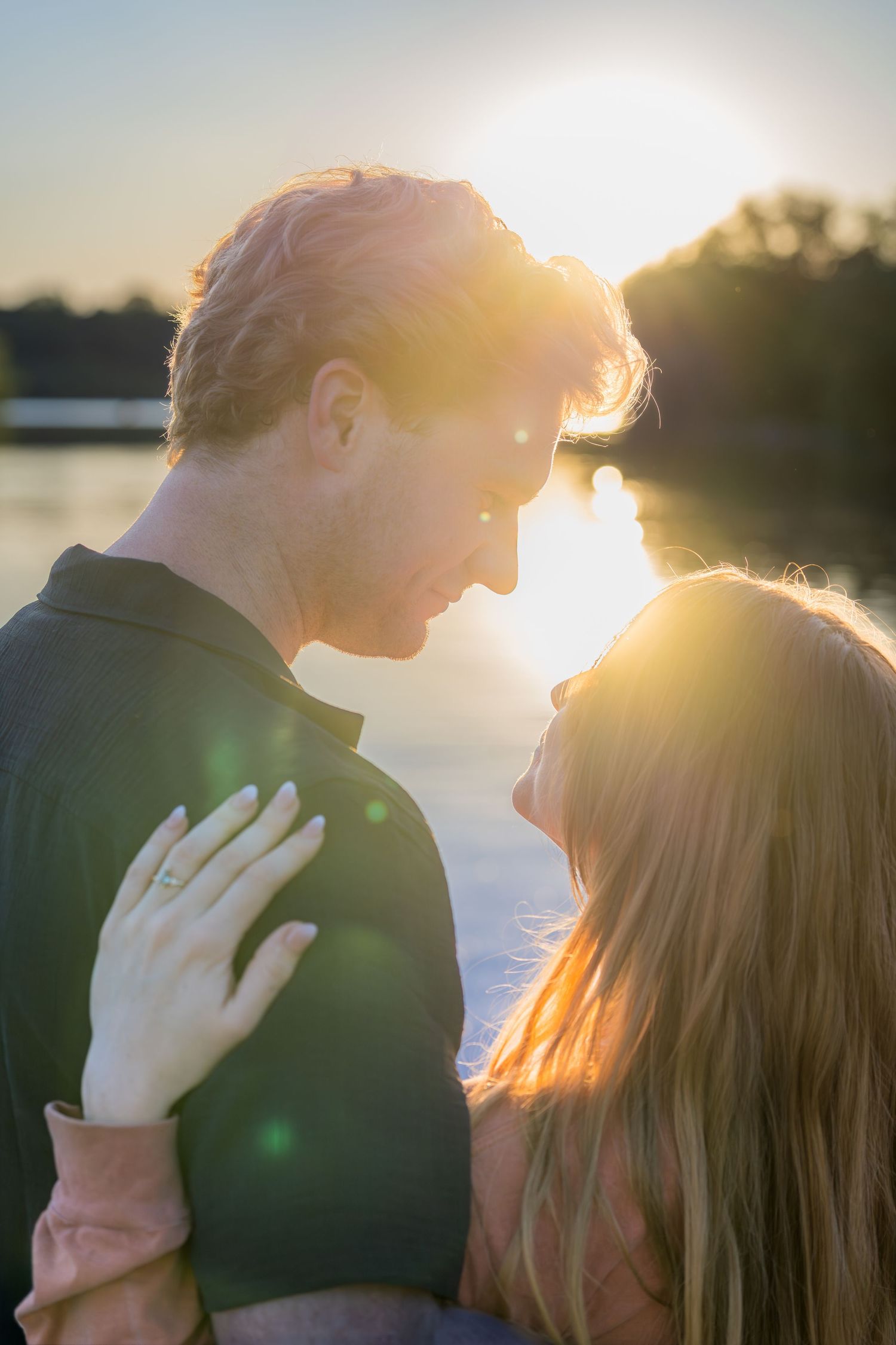 Couple gazing into each others eyes at sunset near a lake, woman showing an engagement ring.