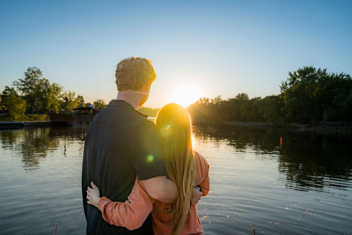 A couple embracing and watching a beautiful golden sunset over a tranquil lake surrounded by trees.