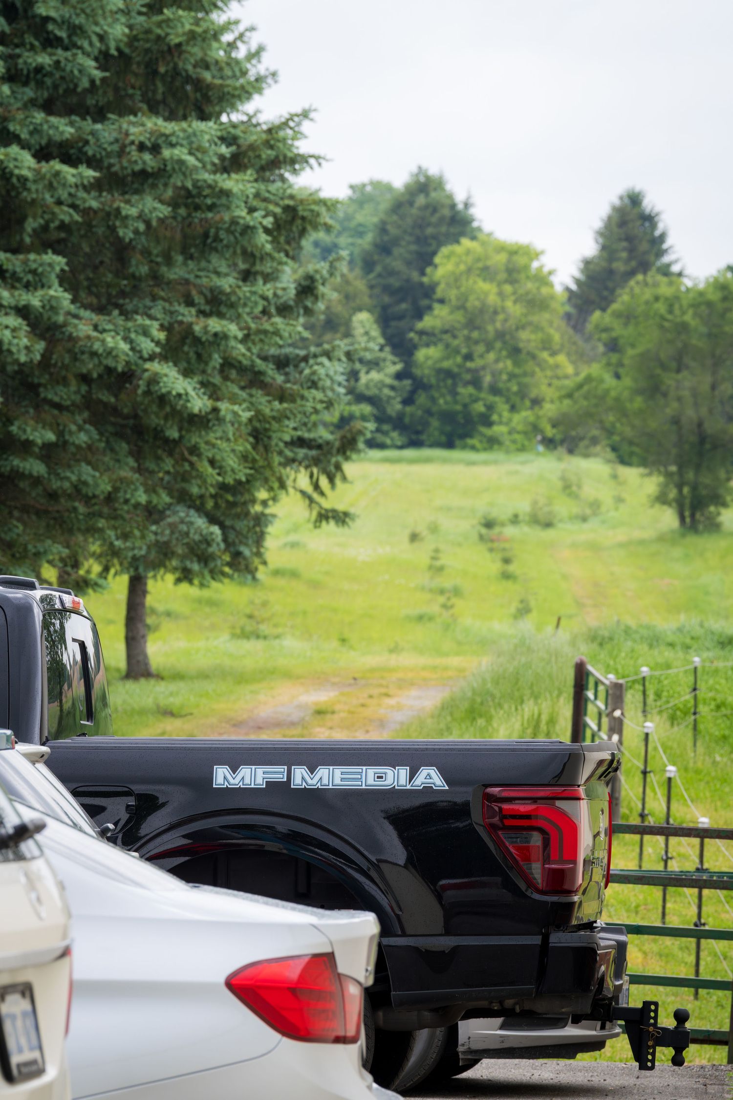 Black MF Media pickup truck tailgate parked in a rural setting with lush green fields and trees in the background.