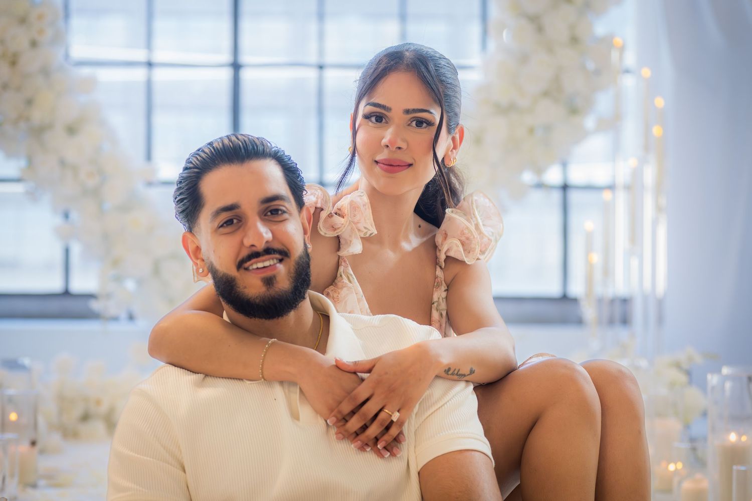 Smiling couple posing together with white floral and candle romantic backdrop behind them.