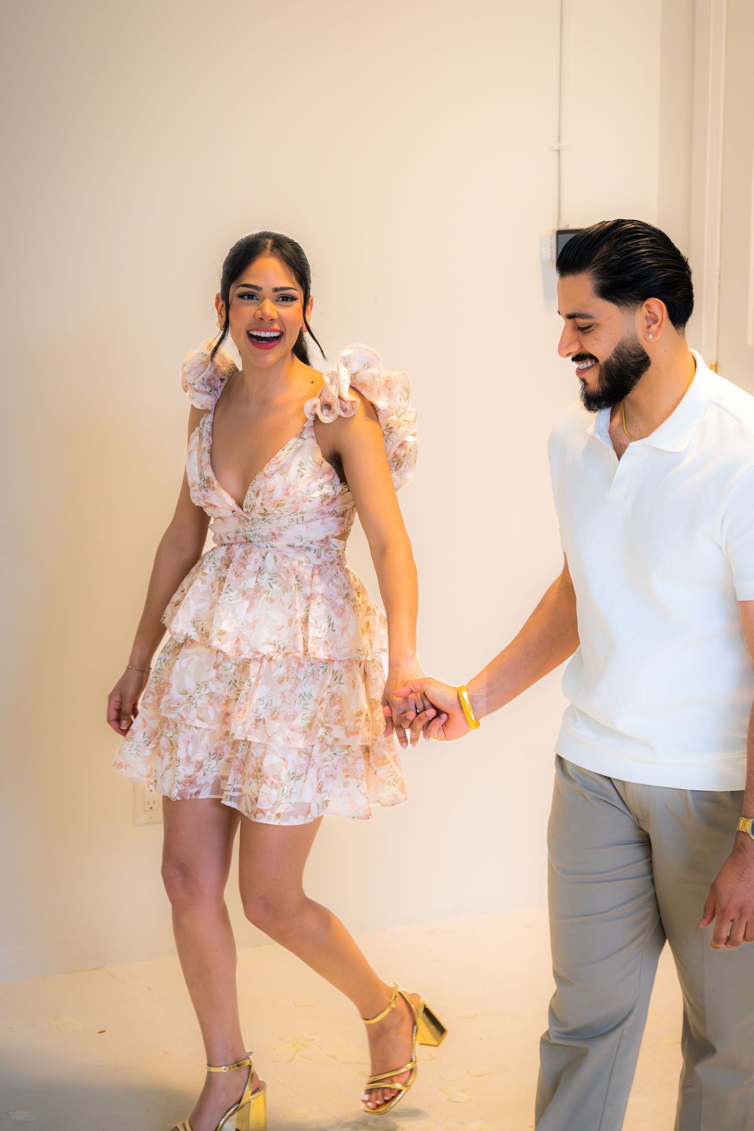 Laughing couple holding hands and walking together, woman wearing a ruffled floral mini dress with gold heels.