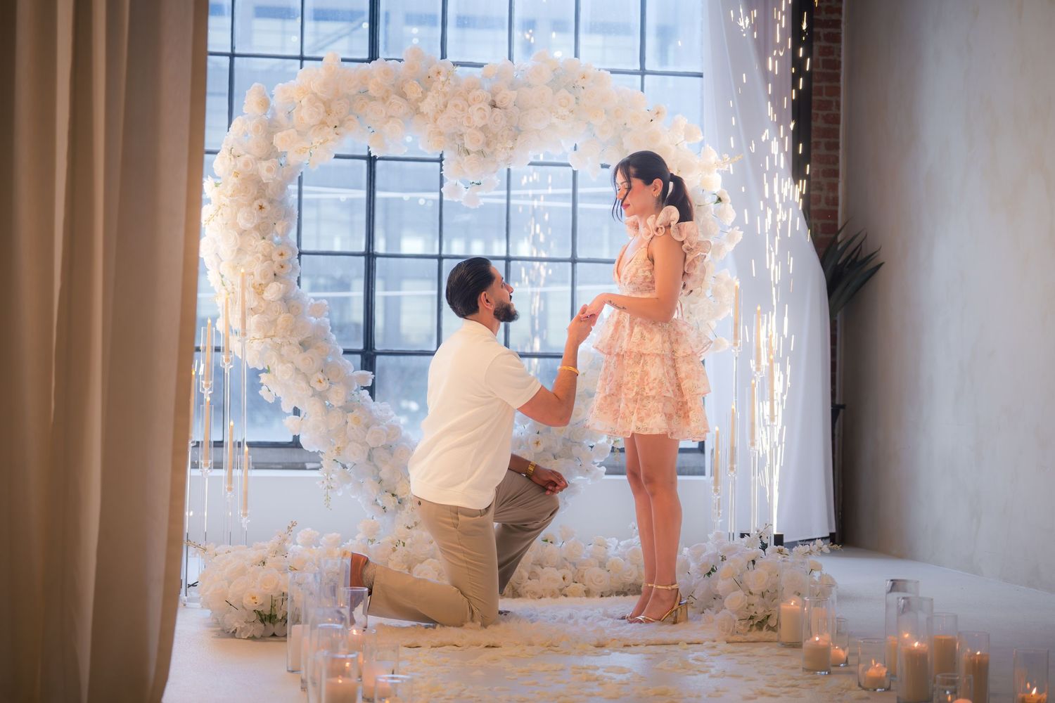 Man kneeling proposing to woman inside heart-shaped floral arch with candles and sparklers.
