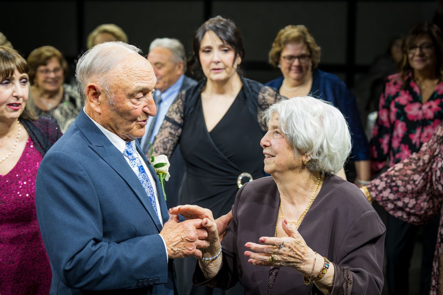 Elderly man in blue suit holding hands with older woman while guests mingle at a festive celebration.