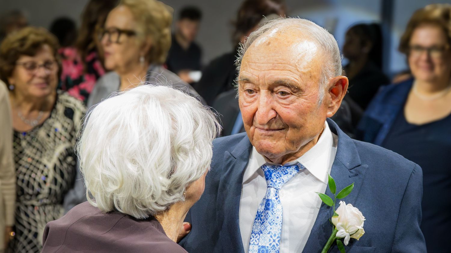 Elderly man in navy suit with boutonniere smiling warmly at a woman during a formal celebration.