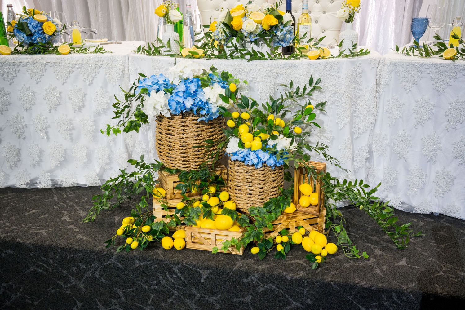 Wicker baskets filled with blue hydrangeas and yellow flowers surrounded by lemons and greenery at an anniversary celebration.