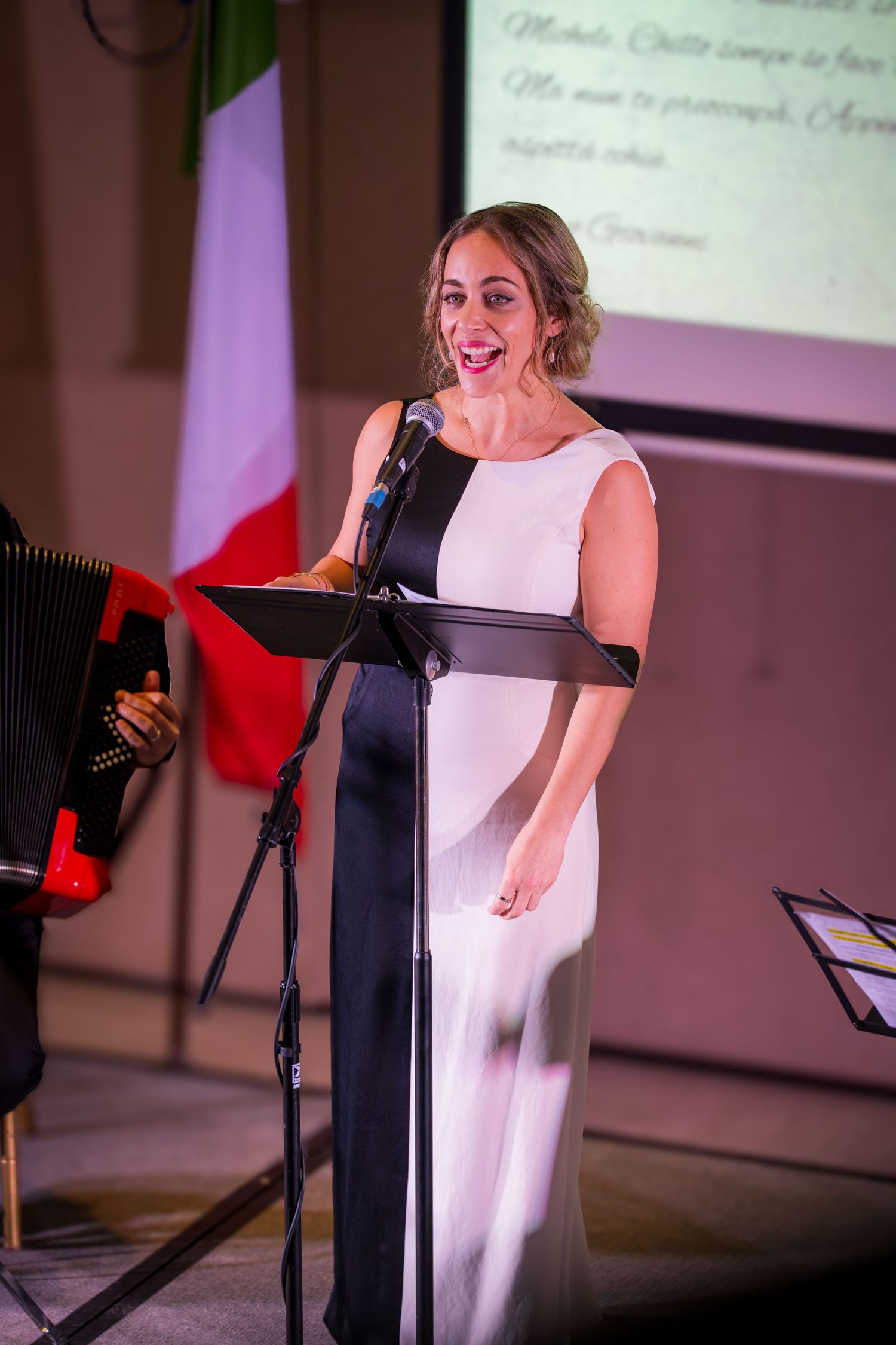 A woman in a black and white dress sings at a podium with an Italian flag and projection screen behind her.