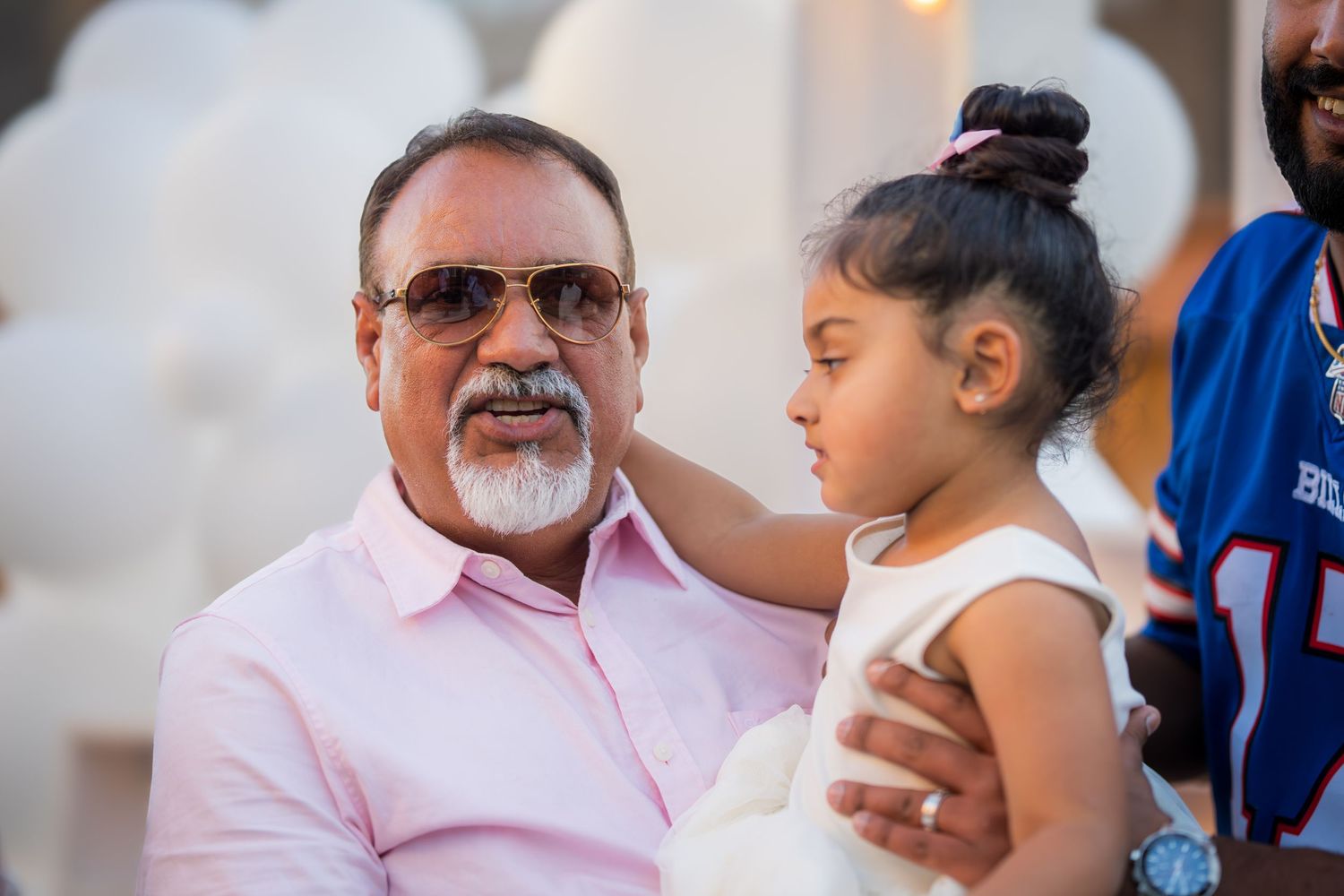A grandfather holds his young granddaughter in a white dress at an outdoor gender reveal party.
