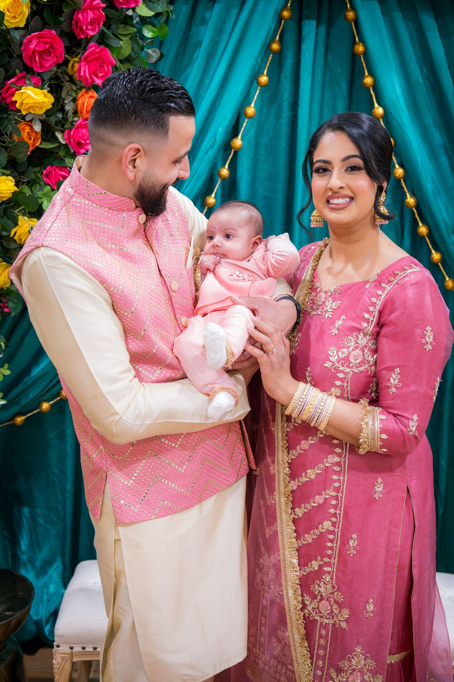 South Asian family in pink traditional attire holding newborn baby at decorated celebration.
