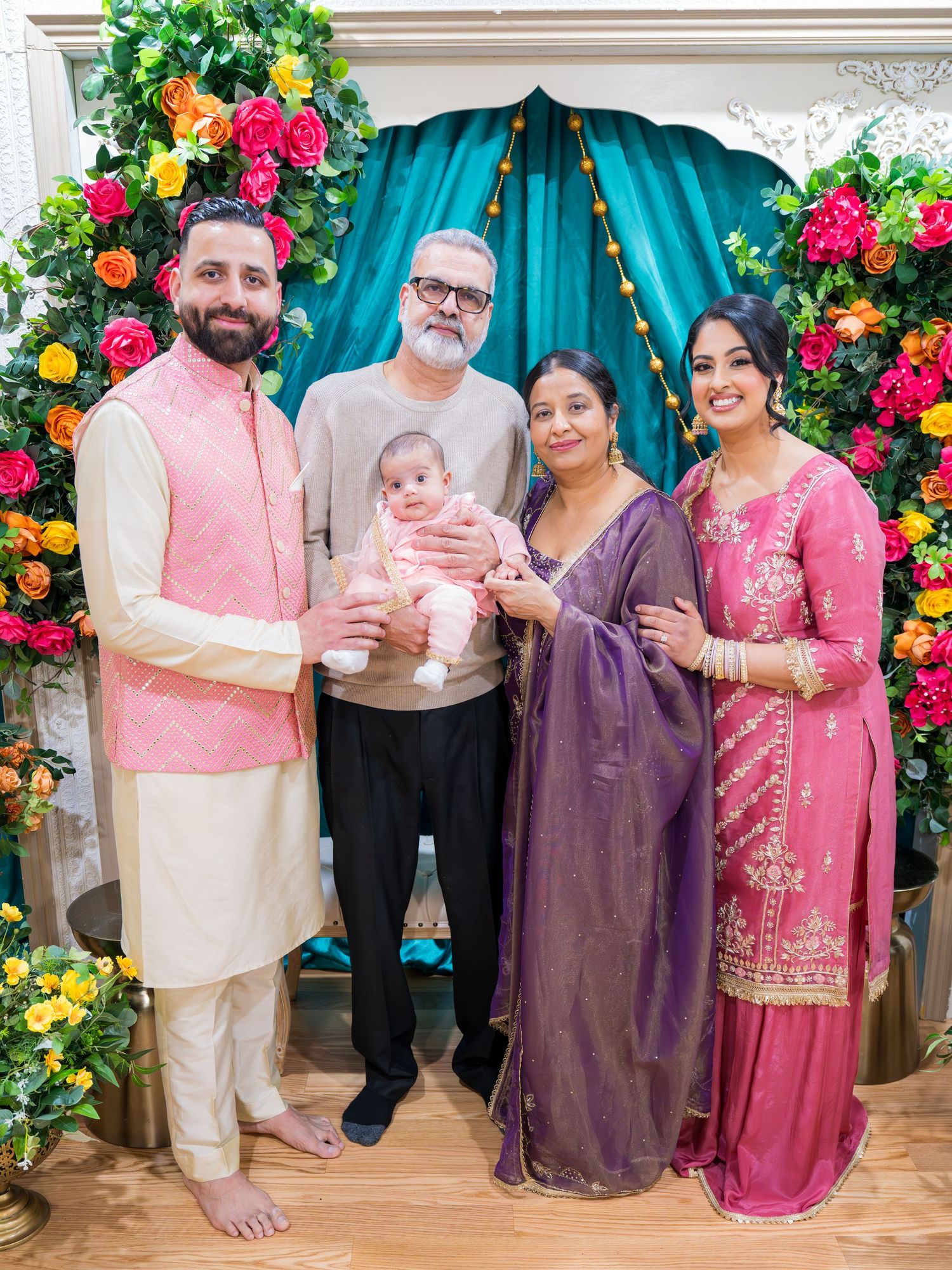 Indian family of five in traditional attire posing with newborn before colorful floral teal backdrop.