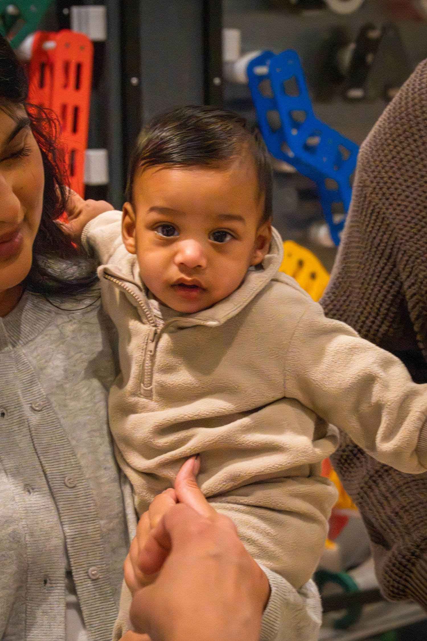 Baby in beige outfit being held by adults near colorful toys.
