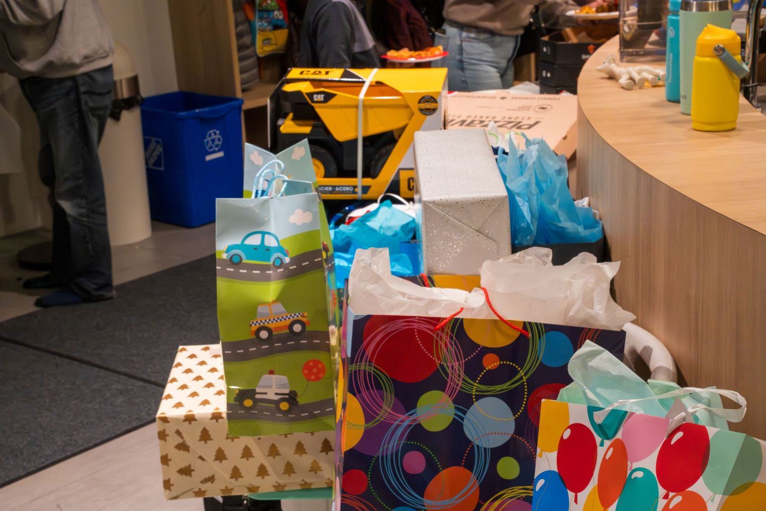 Colorful gift bags and wrapped presents piled on floor at a children's birthday party with toys in background.