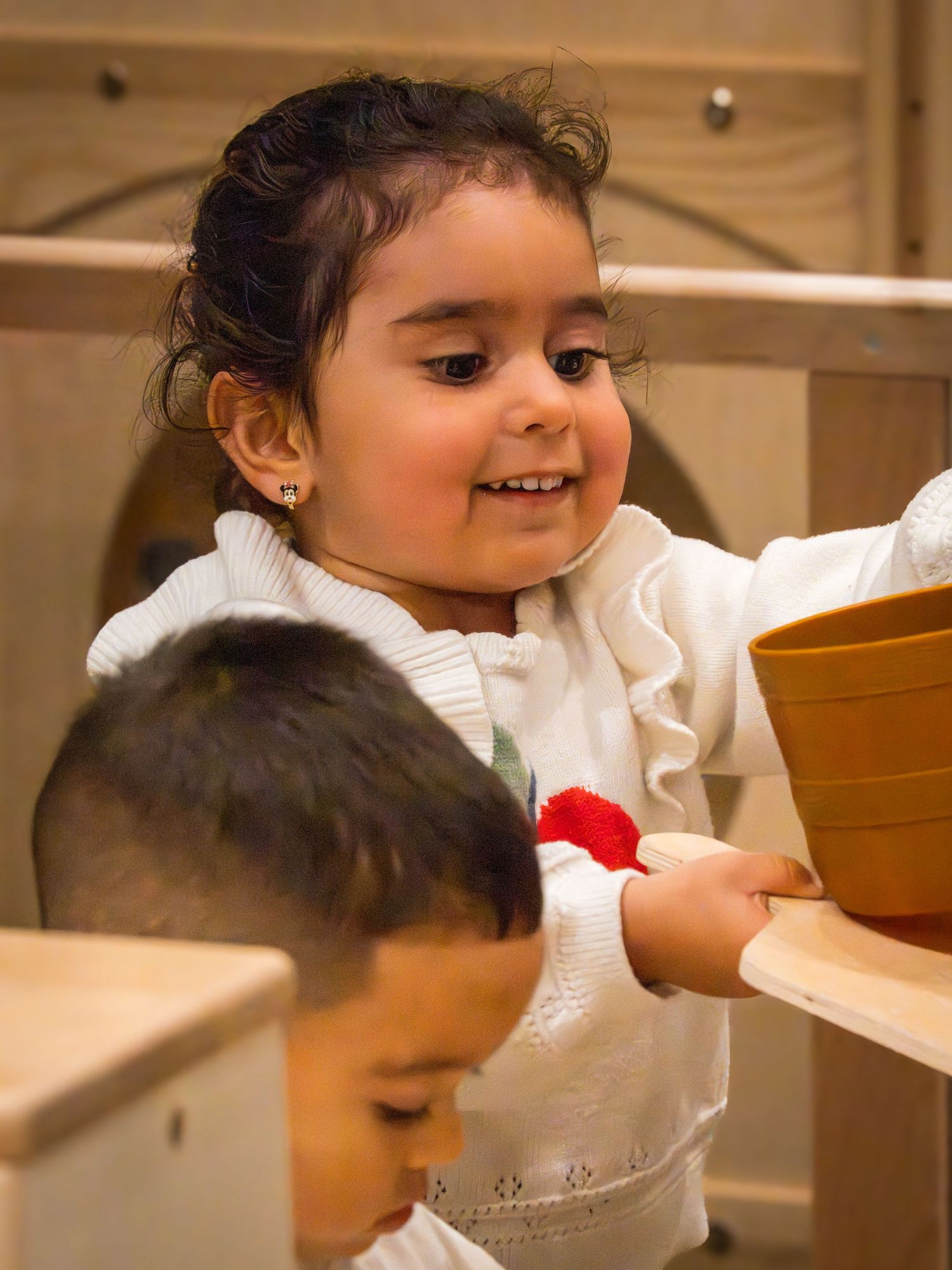 Toddler girl in white apple sweater smiling while holding a wooden bowl at a play area.