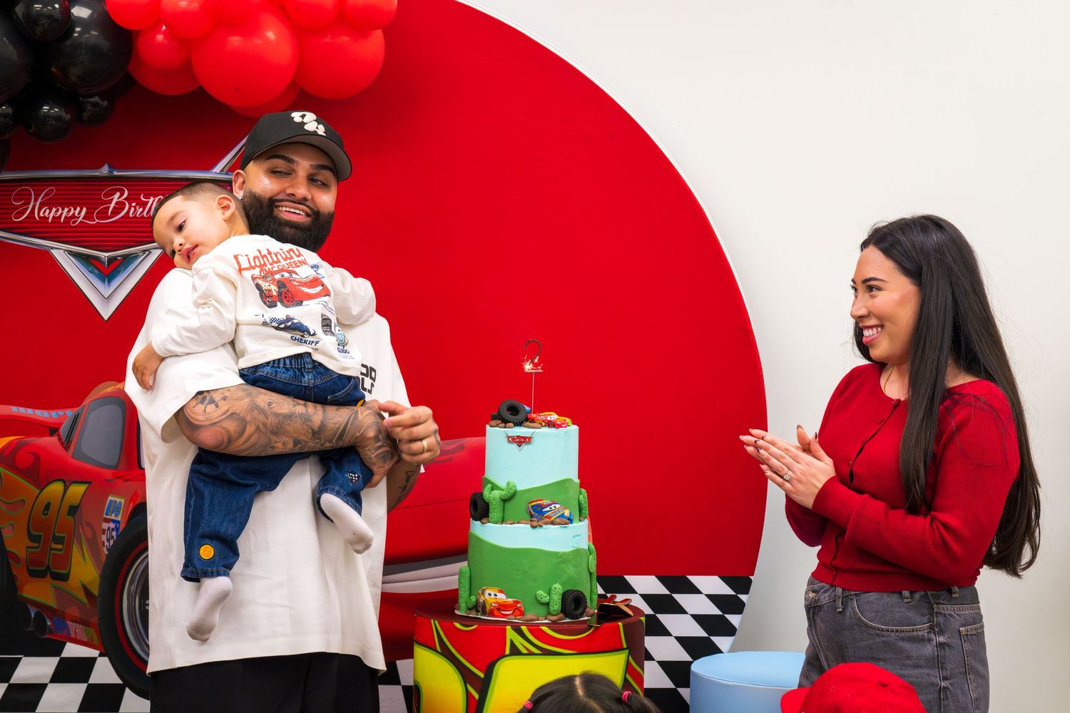 Man holding baby celebrates birthday with woman beside Cars-themed cake and red decorations.