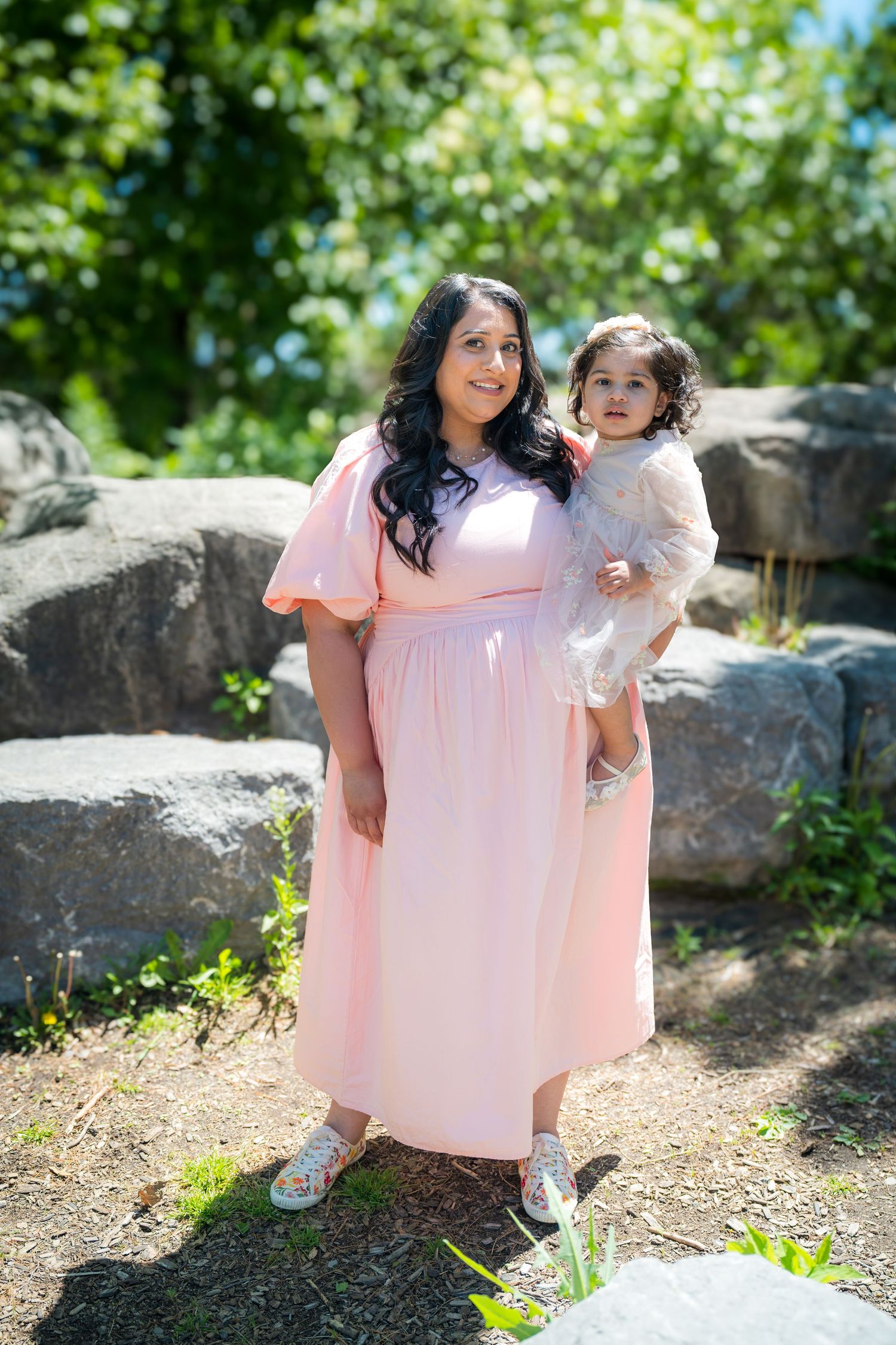 Mother in pink maxi dress holding toddler in white dress outdoors in a garden setting.