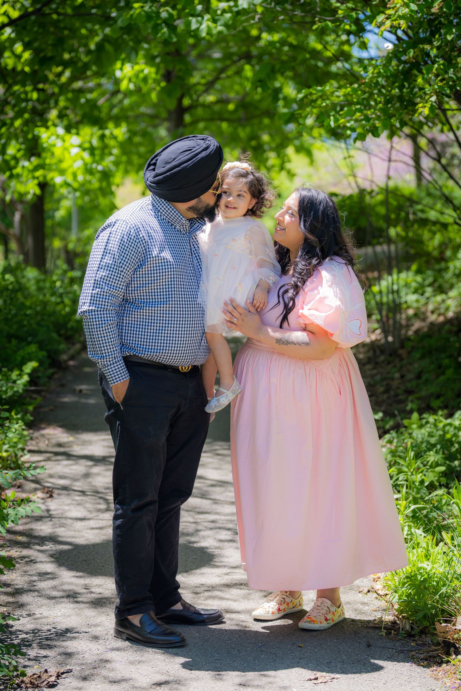 A Sikh family with father in turban holding young daughter while mother in pink dress smiles in lush green park.