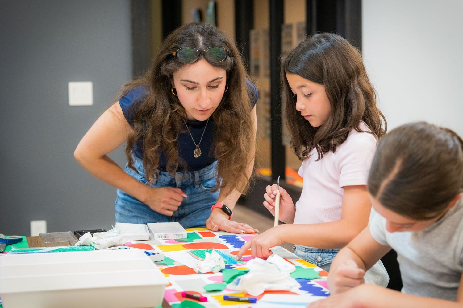 A female teacher helps two young girls with a colorful paper collage art activity.