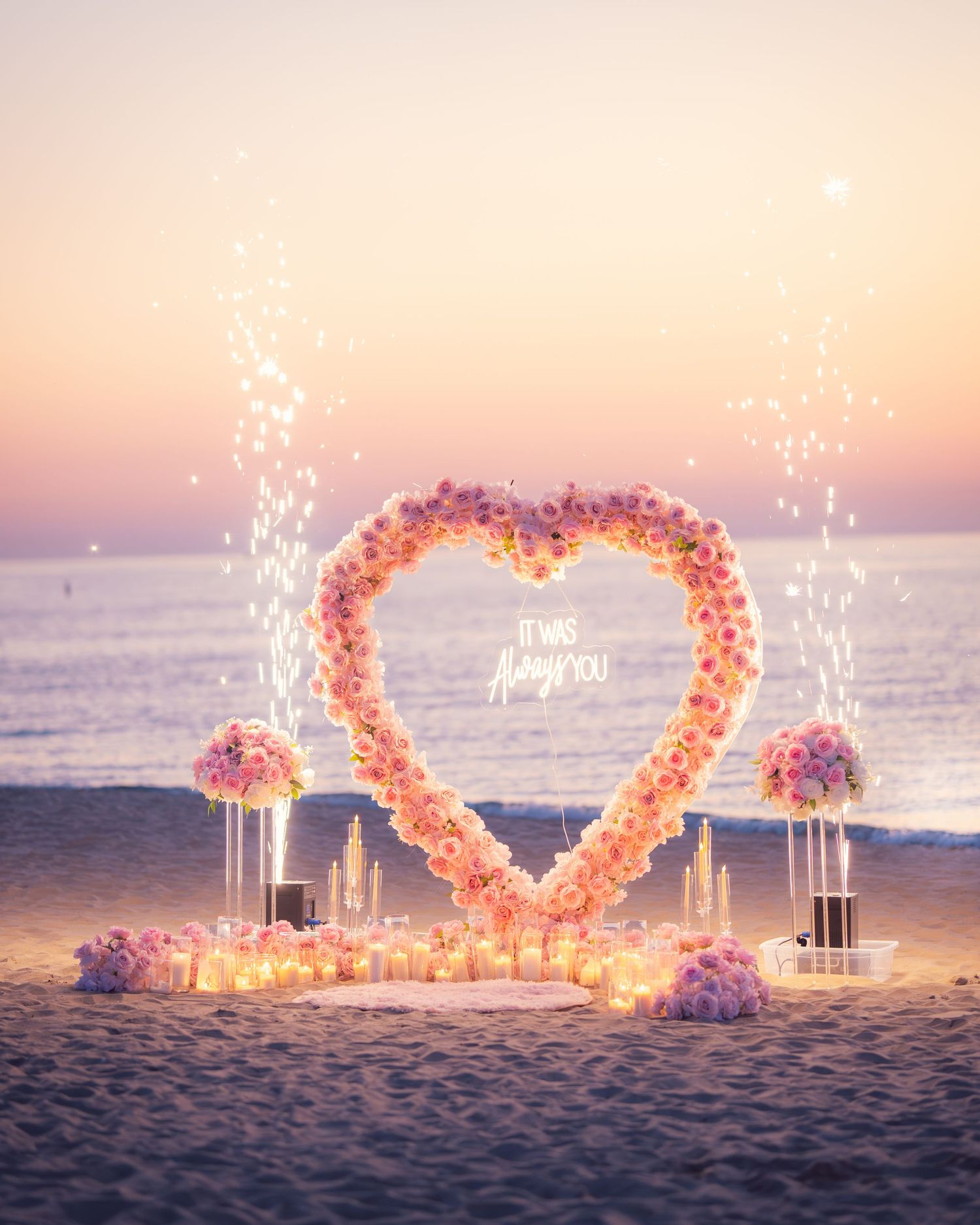 Heart-shaped floral installation with pink roses and candles on beach at dusk.