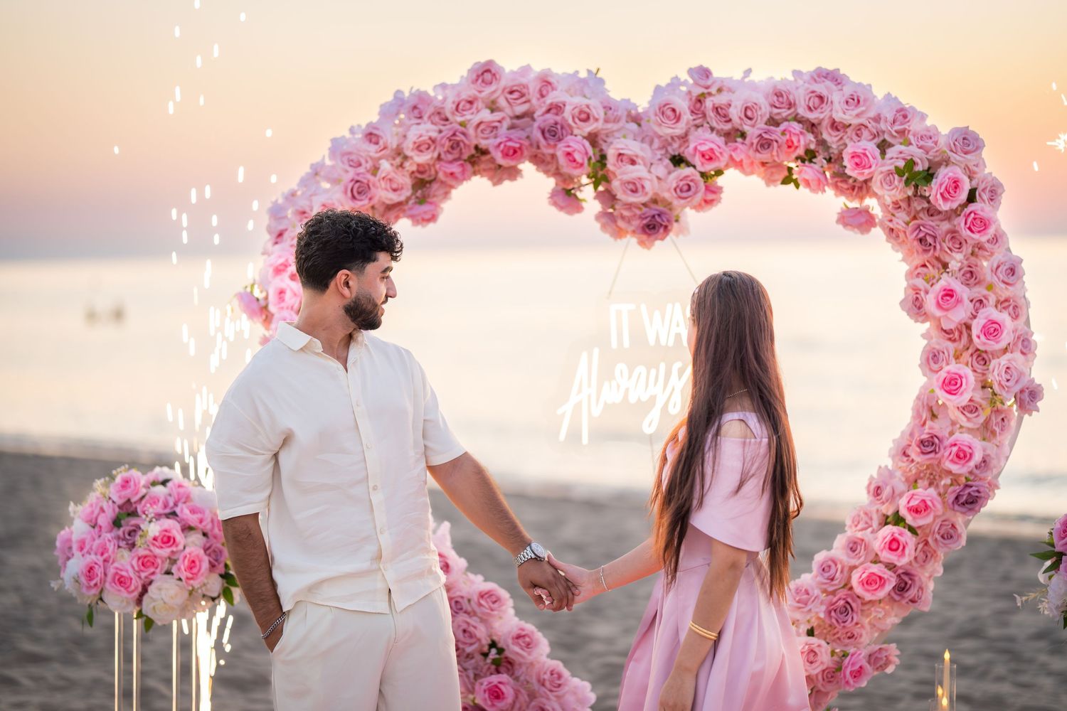 A couple stands beneath a pink floral heart arch on a beach during golden hour.