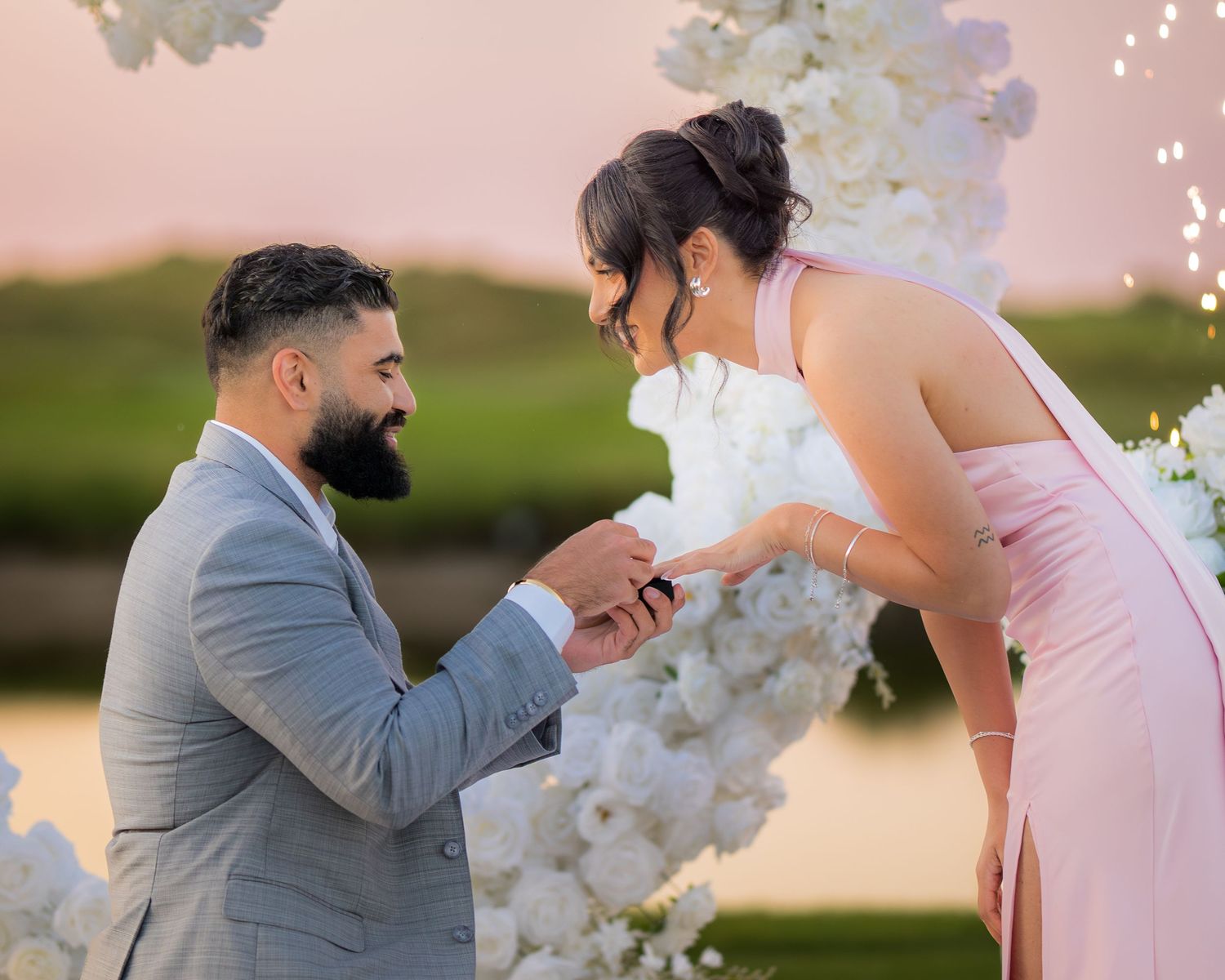 Romantic outdoor proposal scene with white flower arch and glowing candles at sunset.