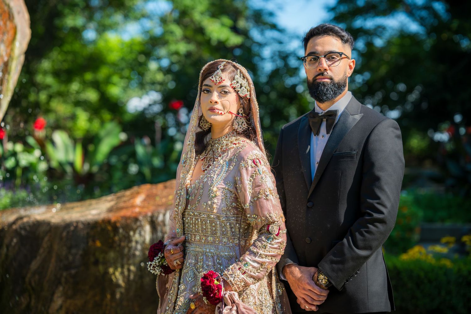 A couple in elegant South Asian wedding attire pose outdoors near a rustic stone wall.