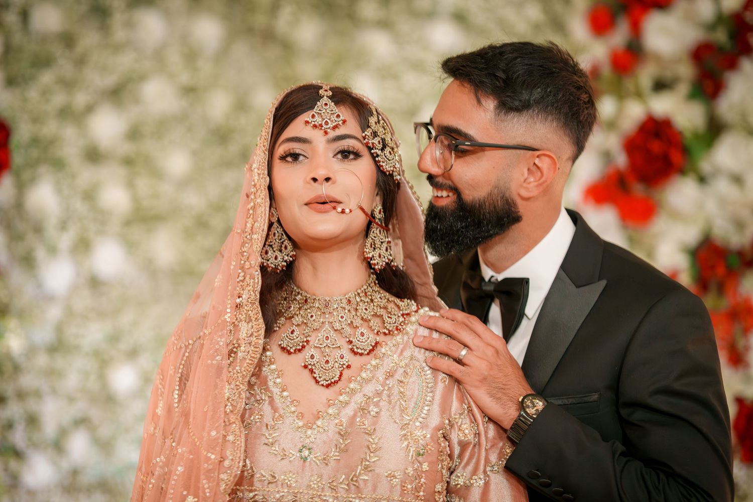 Elegant wedding portrait with couple in traditional South Asian attire against floral wall.