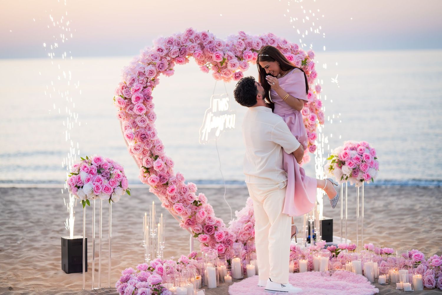 Pink floral heart arch installation on sandy beach during golden hour with ocean backdrop.