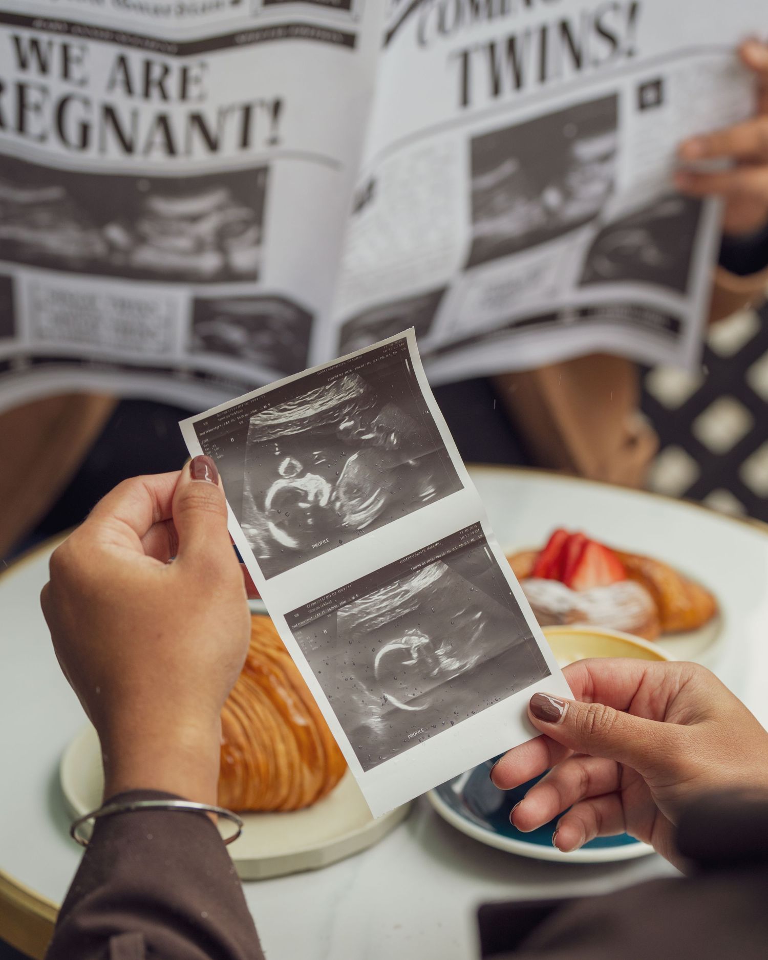 Hands holding ultrasound photos while sitting at cafe table with newspapers and pastries.