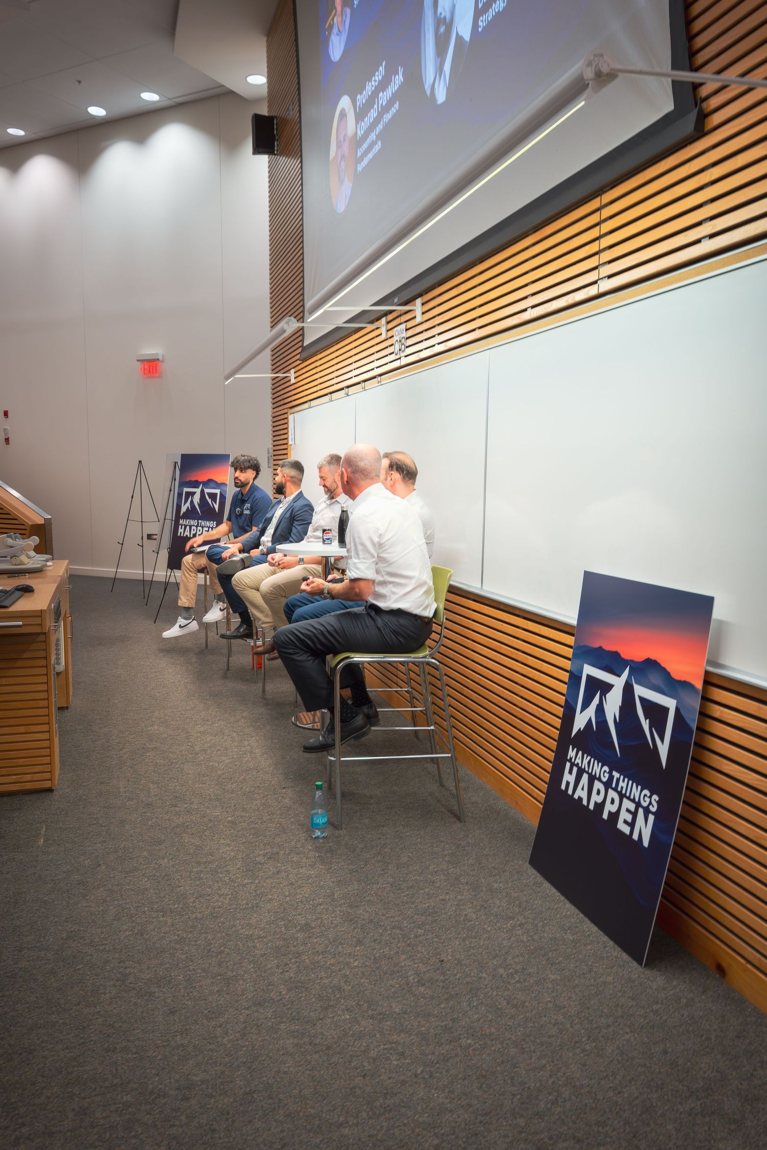 Meeting room with projection screen and wooden wall panels showing attendees seated in chairs.