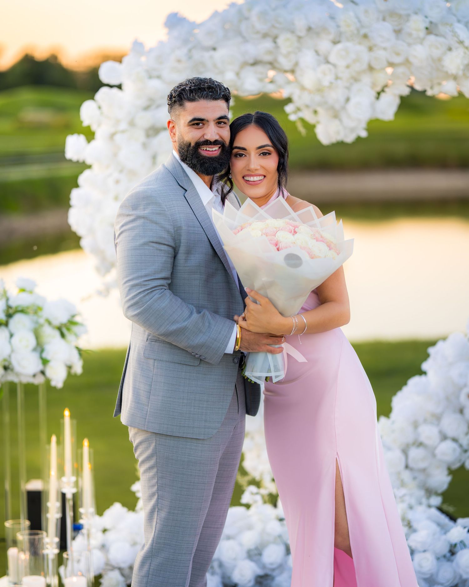 A romantic outdoor proposal scene with white floral arch decorations at sunset.