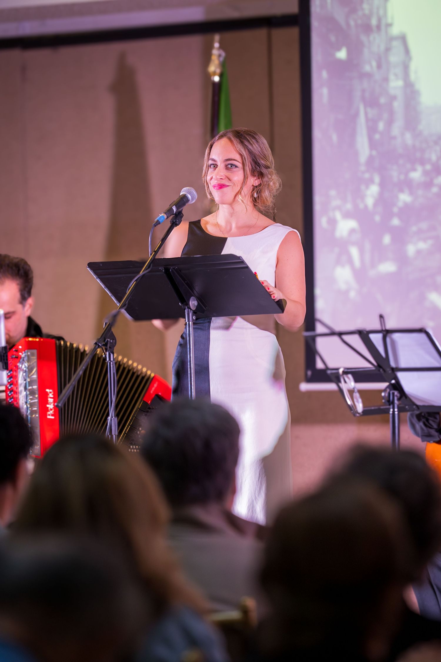 A woman in a white dress performs at a microphone stand during a live music event with an accordionist beside her.
