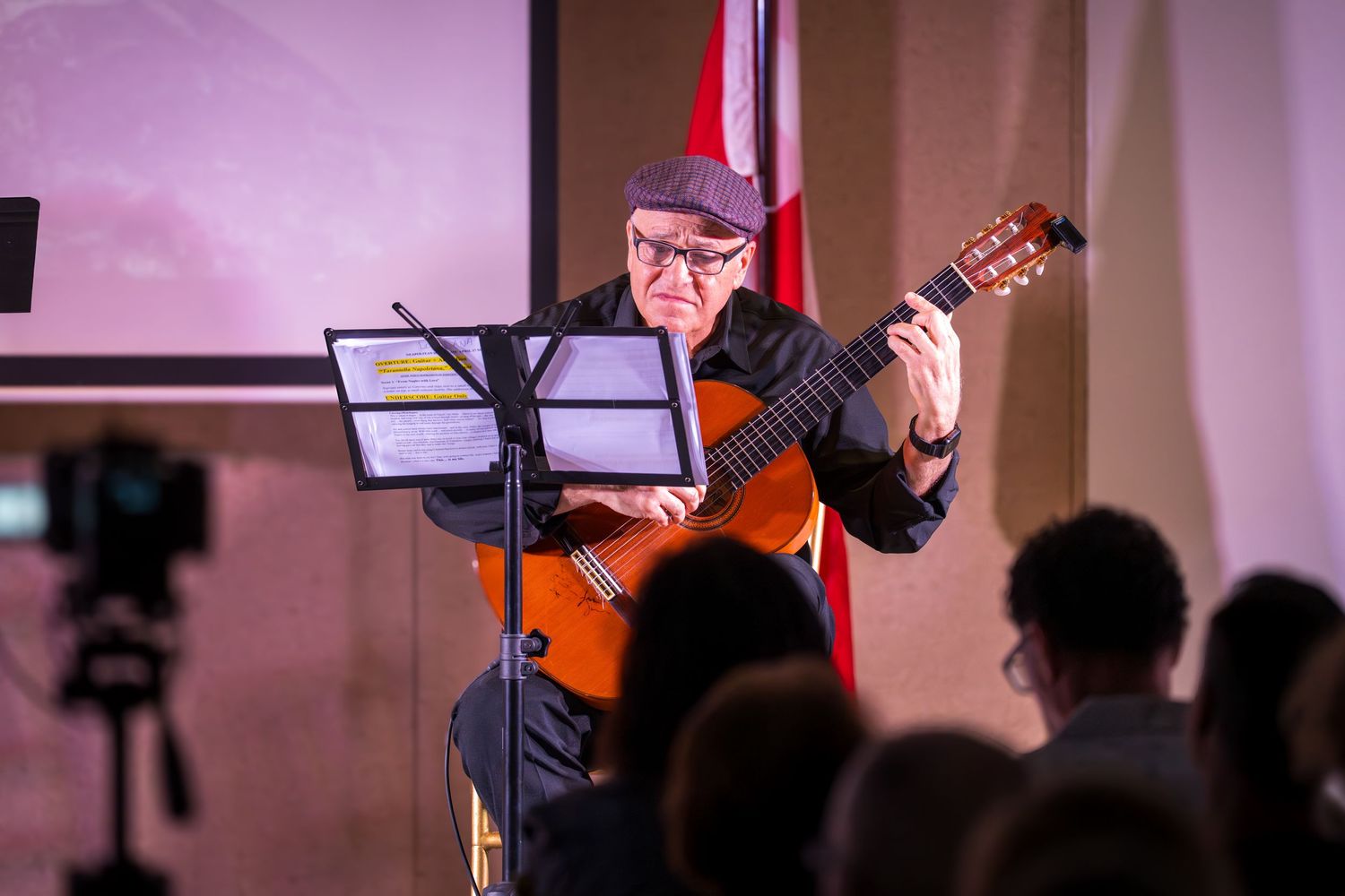 Elderly guitarist in cap plays classical guitar on stage before an audience at an indoor concert.