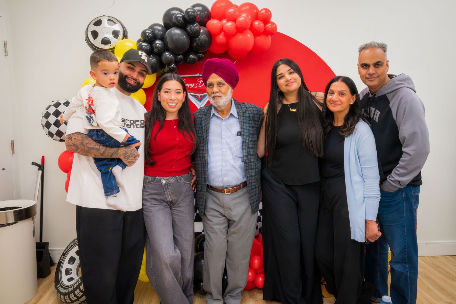 Family group photo of seven people smiling together in front of a red and black balloon arch at a celebration.